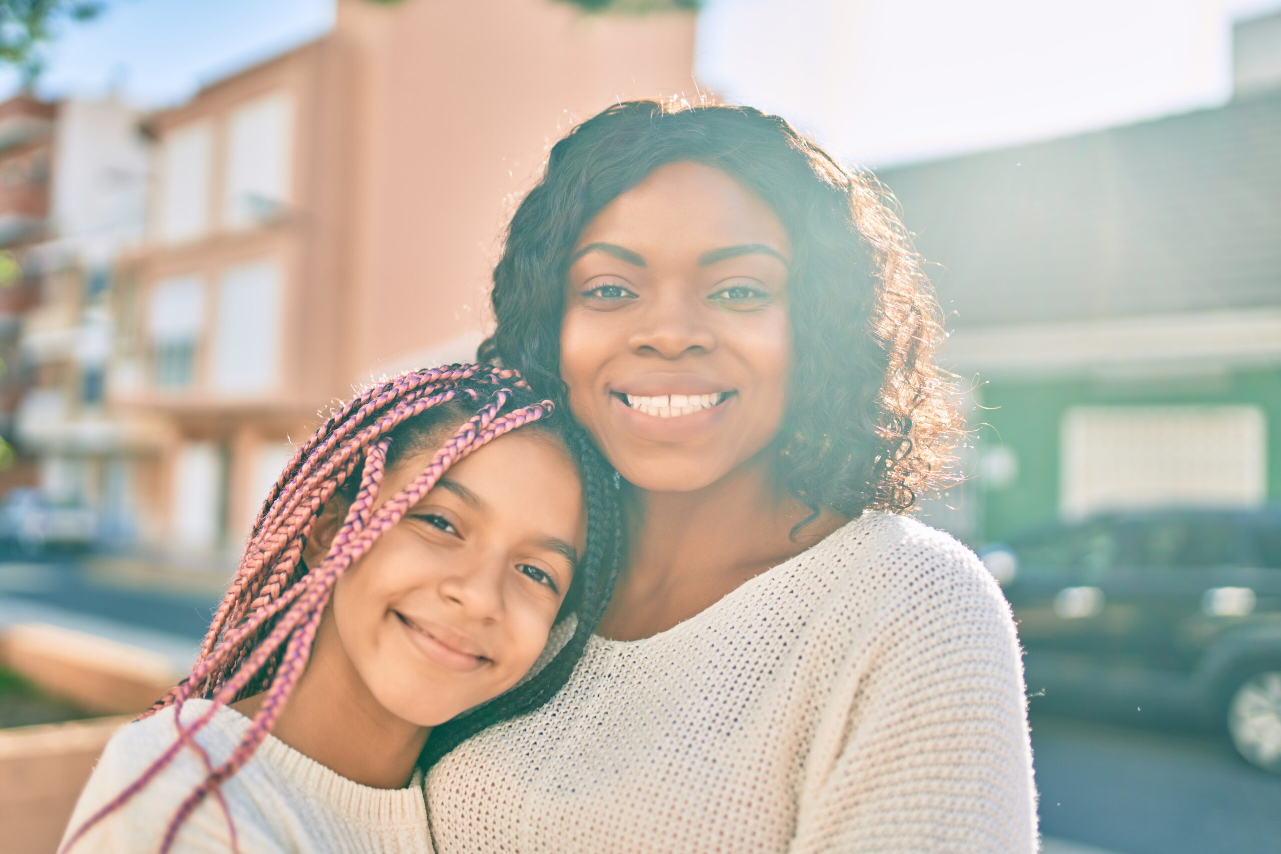A smiling Black mother and daughter stand close together outdoors in soft sunlight, both wearing cream sweaters, exuding warmth, love, and connection.