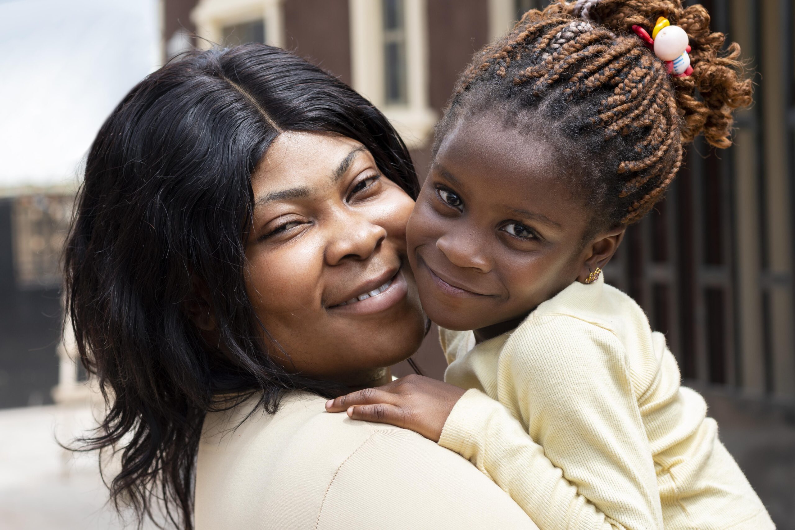 A Black mother holds her daughter close, both smiling softly at the camera, their cheeks touching in a tender moment of affection.