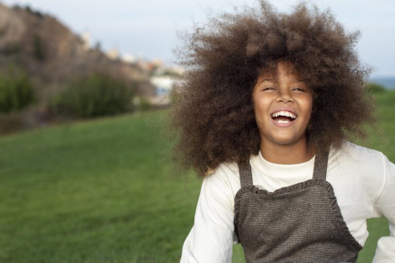 A Black girl with a voluminous afro laughs freely in an open green field, her joy radiant and unfiltered.
