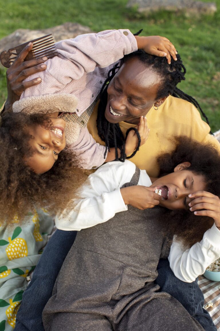 A joyful Black parent with locs playfully embraces two young children with curly hair, all laughing together in a moment of warmth and connection on a picnic blanket.