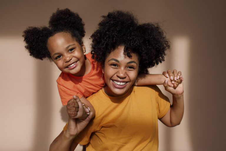 A mother and young daughter with curly hair smile joyfully in a studio setting, dressed in vibrant orange and yellow tones, celebrating their bond.
