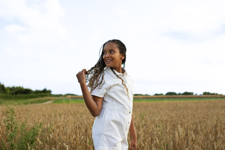 A Black girl with braids smiles while standing in a golden field, wearing a white romper and turning playfully to the side, bathed in soft natural light.